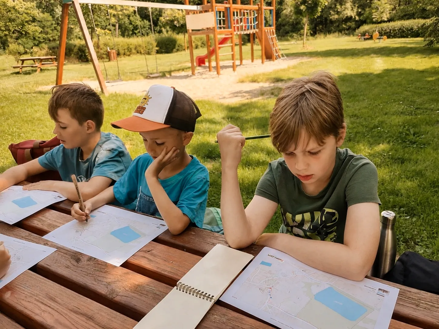 Tři děti pracují venku u stolu s mapami a pracovními listy