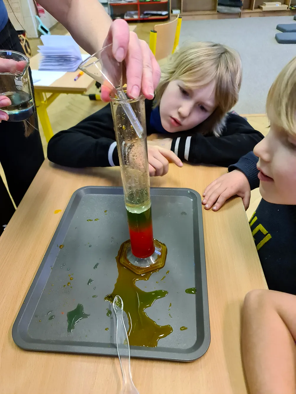 Ruce pedagožky nalévají barevnou kapalinu do válce, dvě děti pokus zblízka sledují